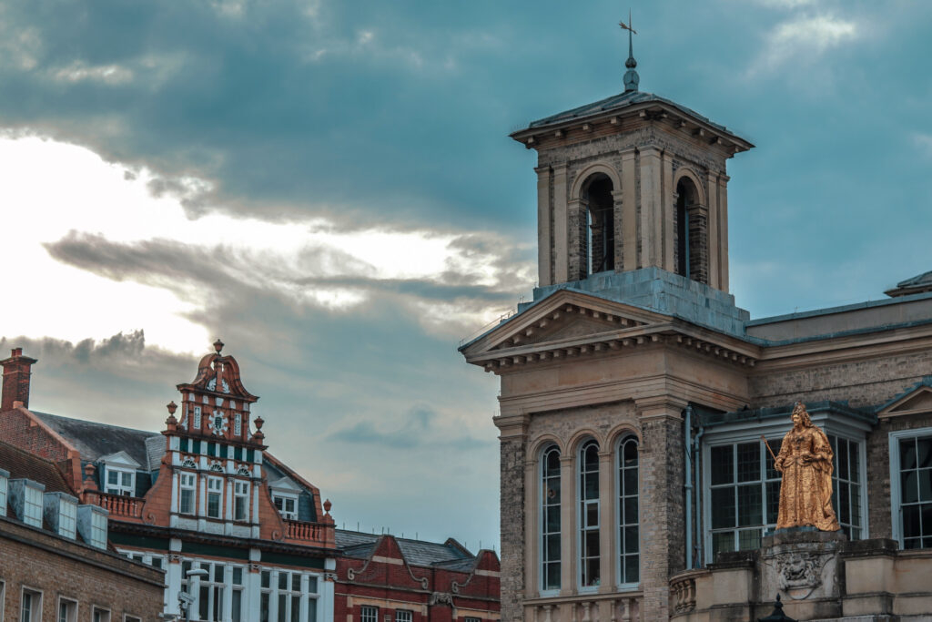 Architectural view of the Market House in Kingston upon Thames, featuring a prominent golden statue of Queen Anne and a square bell tower against a cloudy, dramatic sky.