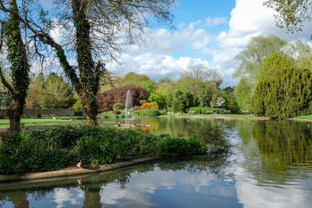 Pinner Memorial Park, Pinner, Middlesex, UK. Photo taken on a sunny (partially cloudy) spring day, showing lake with fountain, trees and green foliage.