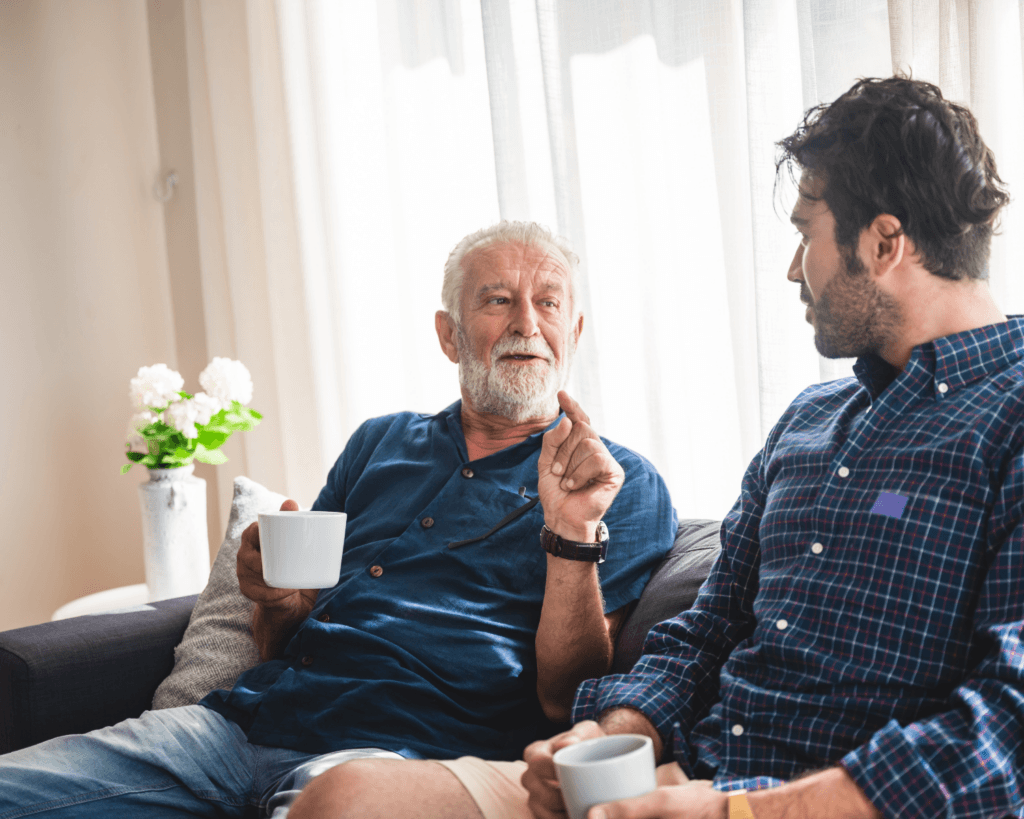 An older gentleman sat indoors on the sofa speaking with a carer. Both are staying hydrated with a drink.