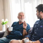 An older gentleman sat indoors on the sofa speaking with a carer. Both are staying hydrated with a drink.