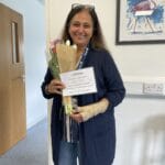Professional carer standing in the Ashridge Home Care office. She is smiling into the camera and holding a bunch of tulips. She is also holding a certificate to celebrate her one year work anniversary.