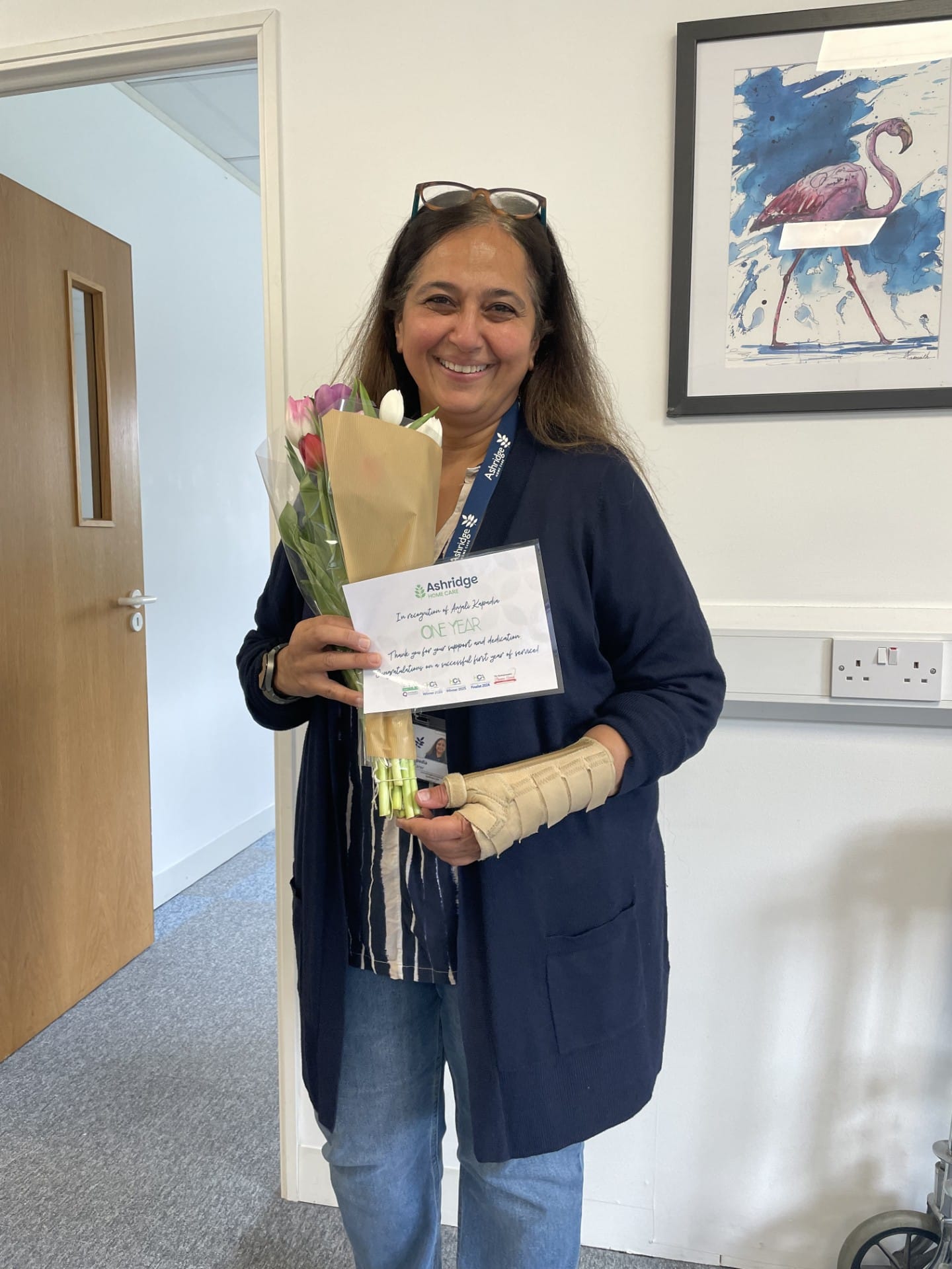 Professional carer standing in the Ashridge Home Care office. She is smiling into the camera and holding a bunch of tulips. She is also holding a certificate to celebrate her one year work anniversary.