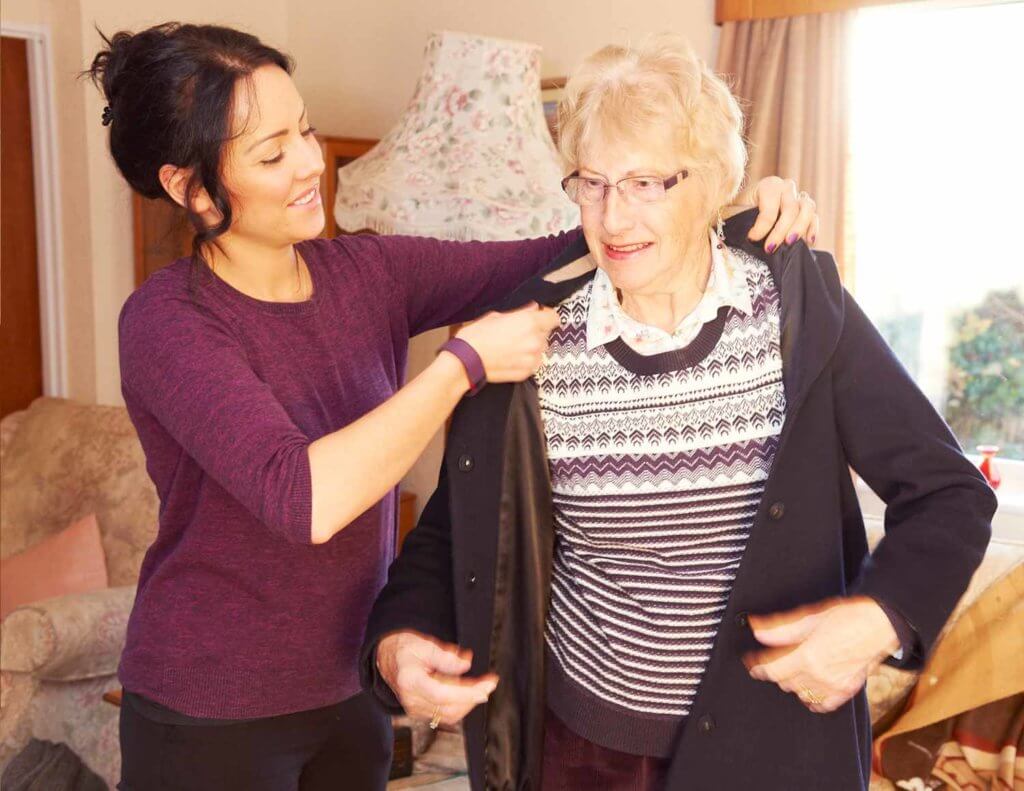 A carer helps an elderly woman put her jacket on